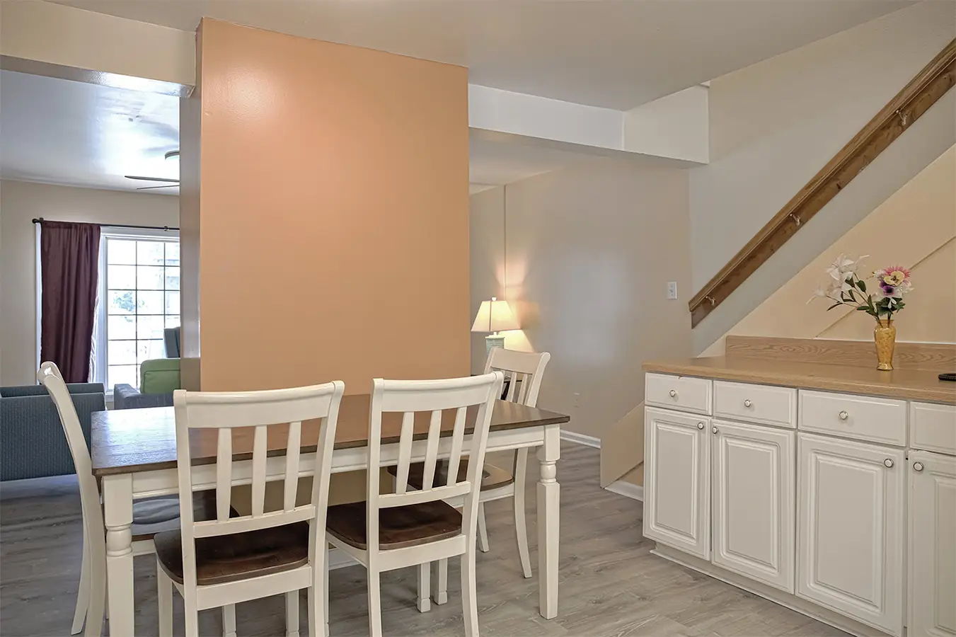 The dining area of an apartment, with a white table and chairs by the window and a staircase behind