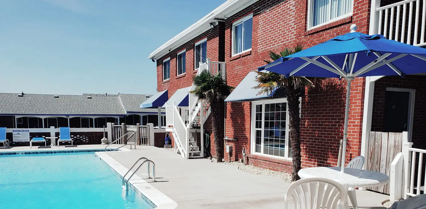 The outdoor pool in front of the motel\u2019s red brick building, with blue awnings, umbrellas and white sun loungers