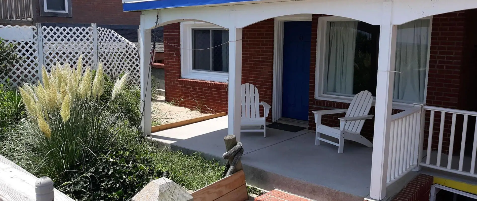 A covered porch with white Adirondack chairs beside a blue room door and pampas grass